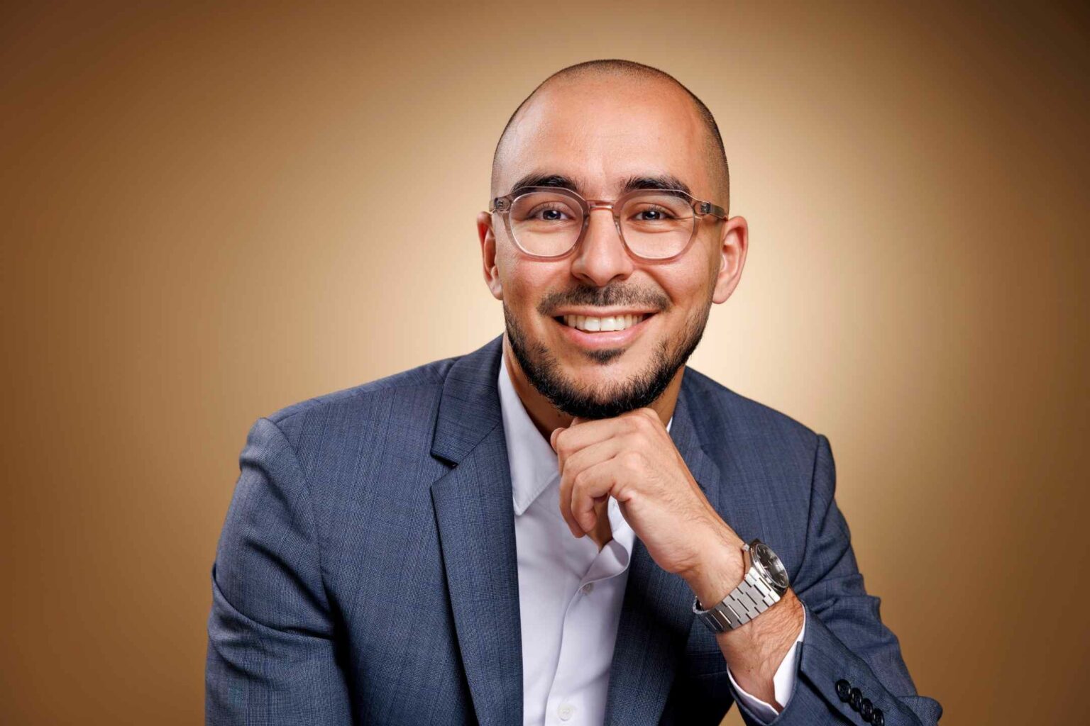 Portrait corporate d’un homme souriant avec lunettes et costume bleu, photographié en studio sur fond brun chaud